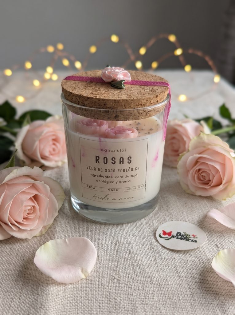 Vela de rosas en vaso de cristal con tapa de corcho, rodeada de rosas y luces navideñas
